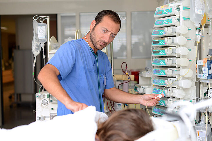 Eine männliche Pflegekraft steht am Patientenbett zwischen vielen Infusionen und Schläuchen. 