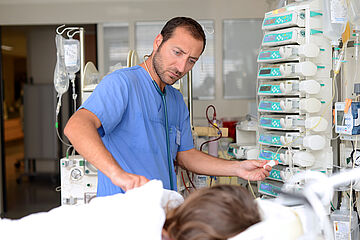 Eine männliche Pflegekraft steht am Patientenbett zwischen vielen Infusionen und Schläuchen. 