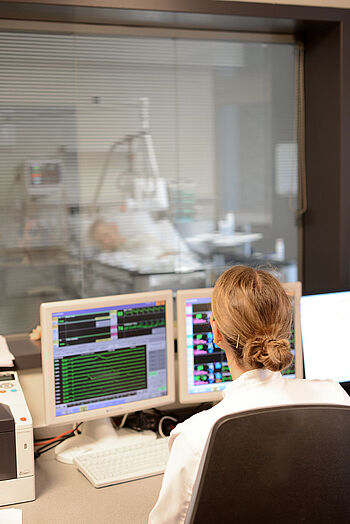 Eine Pflegekraft sitz vor den Monitoren für Monitoring und kann gleichzeitig duch eine Überwachungsfenster einen kritischen Patienten im Blick behalten.